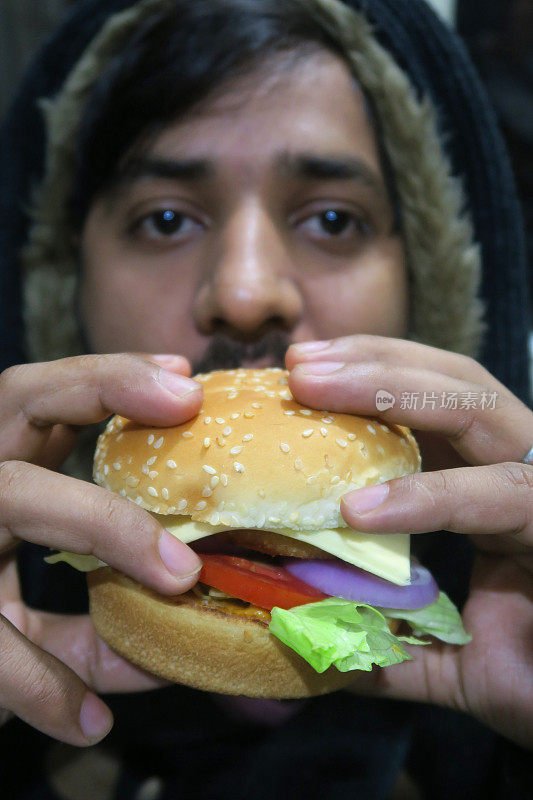 一名印度男子拿着汉堡从前面露出脸，吃着健康的自制奶酪汉堡/汉堡/牛肉汉堡，汉堡包着芝麻籽、融化的奶酪、番茄、红洋葱片和莴苣